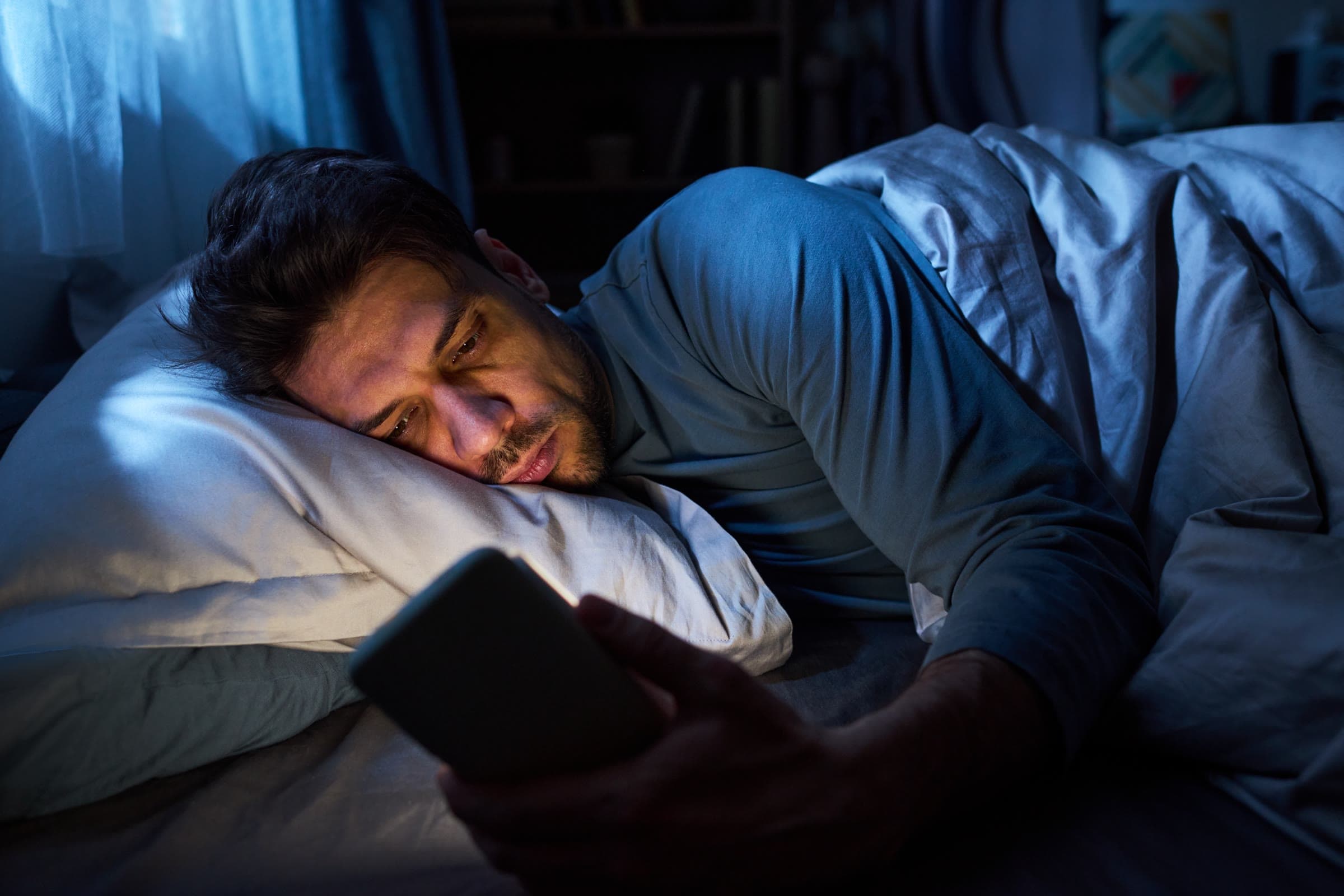 A man lying awake in bed at night, his face lit by the phone he is looking at.